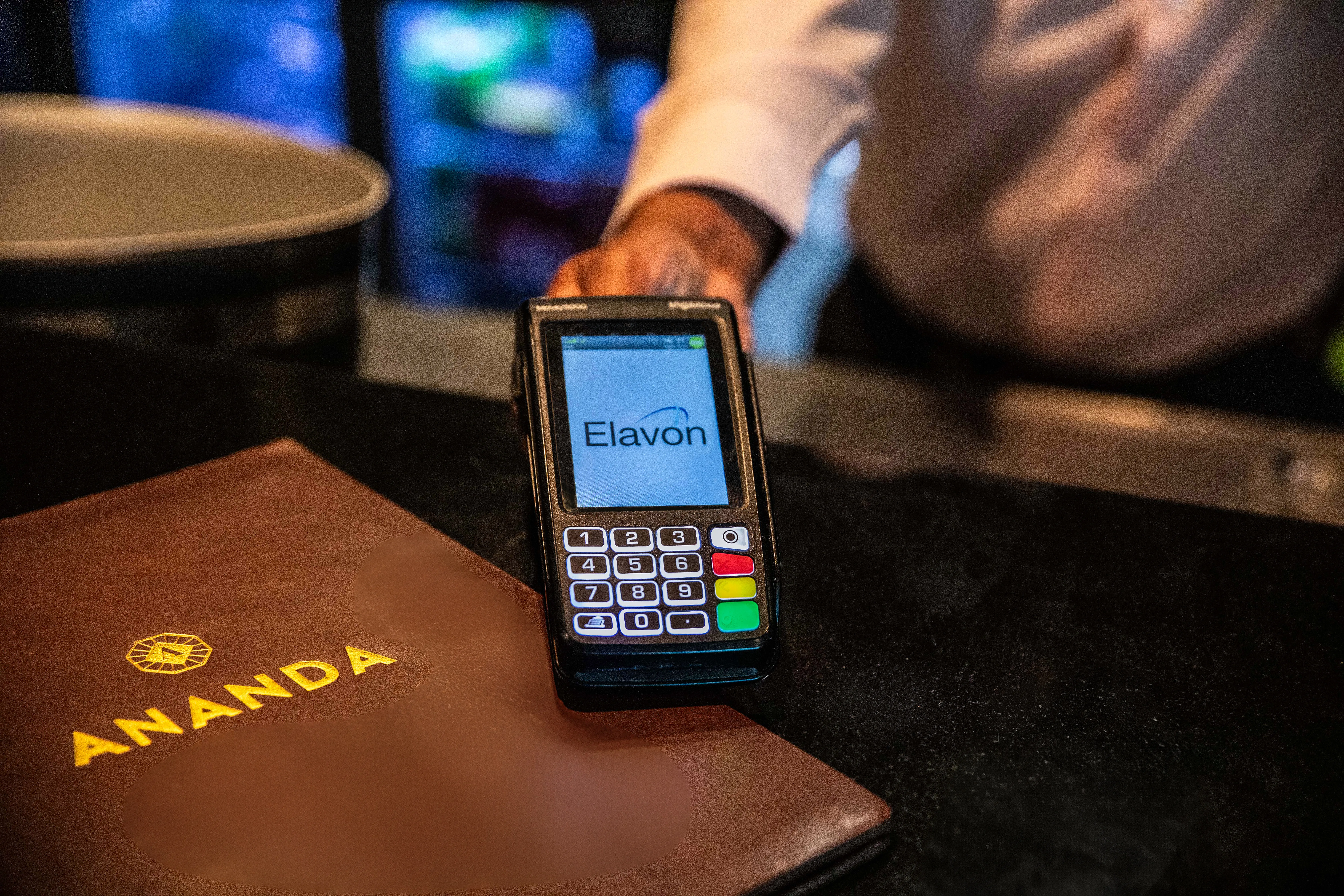 A person in a white shirt holds an Elavon-branded payment terminal, ready for use. Beside the card machine lies the menu for Ananda, part of the Jaipur restaurant group